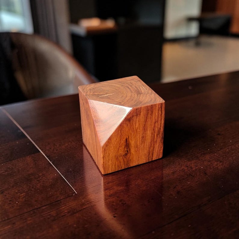 May include: A handcrafted wooden cube with a geometric design. The cube is made of a reddish-brown wood, with a polished finish. One corner is cut at an angle, creating a unique shape. The cube is displayed on a dark brown surface.
