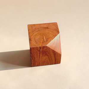 May include: A geometric wooden sculpture with a rich, reddish-brown hue. The block-shaped piece features a diagonal cut on one side, revealing a lighter wood grain. The sculpture is a unique decorative item.