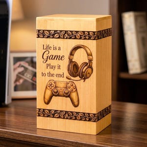 May include: A rectangular light-colored wooden box featuring a laser-engraved design of gaming headphones and a controller. The text reads, Life is a Game Play it to the end. The top and bottom edges are decorated with a detailed leaf pattern border.