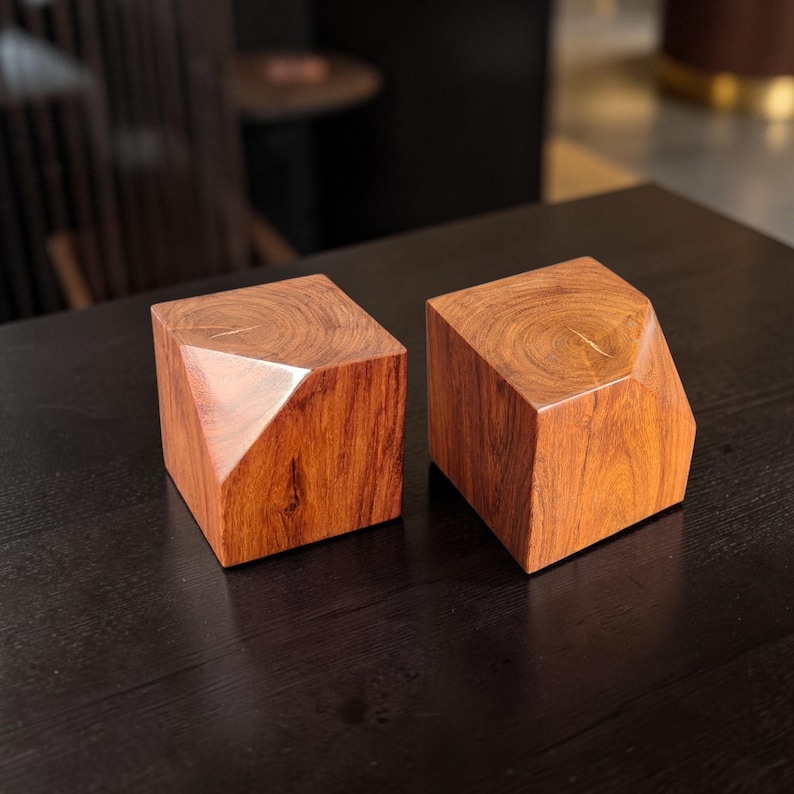 May include: Two geometric wooden blocks with a reddish-brown hue. Each block has a faceted design on one side, creating a unique shape. The wood grain is visible, adding to the natural aesthetic. These decorative blocks could be used as bookends or decorative objects.