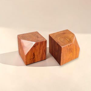 May include: Two geometric wooden blocks with a rich, reddish-brown hue. Each block has a faceted design on one side, creating a unique shape. The wood grain is visible, adding texture and warmth to the objects.