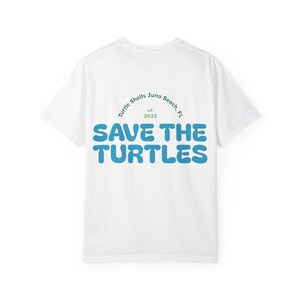May include: White t-shirt with the text "SAVE THE TURTLES" in large, blue, bubble letters. Above the text is a green arched design with the words "Turtle Shells Juno Beach, FL" and the year "2025".