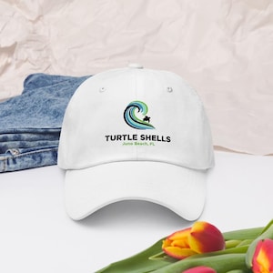 May include: White baseball cap with the text "TURTLE SHELLS" and a wave logo with a turtle silhouette. The text "Juno Beach, FL" is below the logo. The cap is on a white surface.