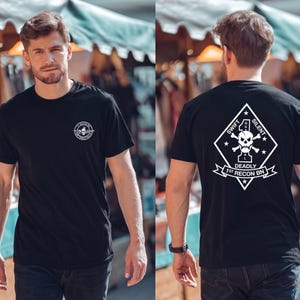 Pode incluir: Camiseta preta com um gráfico branco na frente e atrás. O gráfico frontal é uma caveira e texto em um círculo. O gráfico traseiro é uma caveira e ossos cruzados dentro de um diamante com as palavras "Swift, Silent, Deadly, 1st Recon Bn."