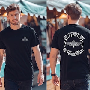 Puede incluir: Camiseta negra con un emblema blanco en la parte delantera y un diseño más grande en la parte trasera. El diseño trasero incluye el texto "U.S. NAVY" y "THE LAST TO LET YOU DOWN" alrededor de un diseño de paracaídas y alas. También se incluye el texto "EST. 1942 PARACHUTE RIGGER".