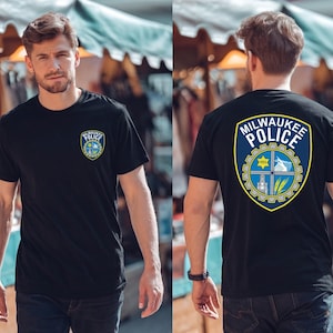 Może przedstawiać: Czarny t-shirt z emblematem Departamentu Policji w Milwaukee z przodu i z tyłu. Emblemat to niebieska tarcza ze złotą obwódką, zawierająca tekst i symbole. Osoba nosi koszulkę na zewnątrz.