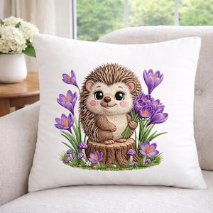 May include: White square cushion featuring an embroidered design of a charming hedgehog clutching purple flowers. The hedgehog is brown with pink cheeks, sitting on a tree stump surrounded by purple crocuses and mushrooms. The cushion is on a white sofa.