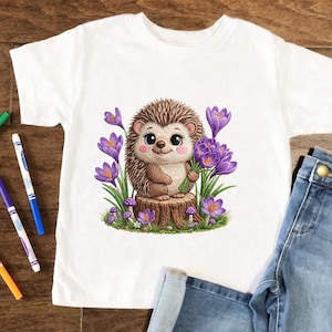May include: White t-shirt with a cartoon hedgehog holding purple flowers, sat on a tree stump. The hedgehog has brown fur and a cute expression. Purple crocus flowers and mushrooms surround the hedgehog.