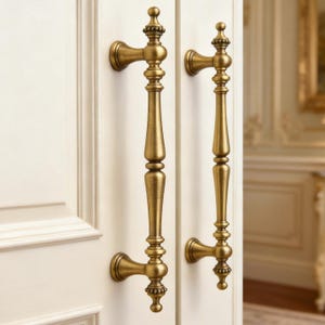 May include: Two antique brass door handles with ornate detailing. The handles are mounted on a white door, showcasing a classic design. The handles have a vertical bar with decorative elements and a curved base.