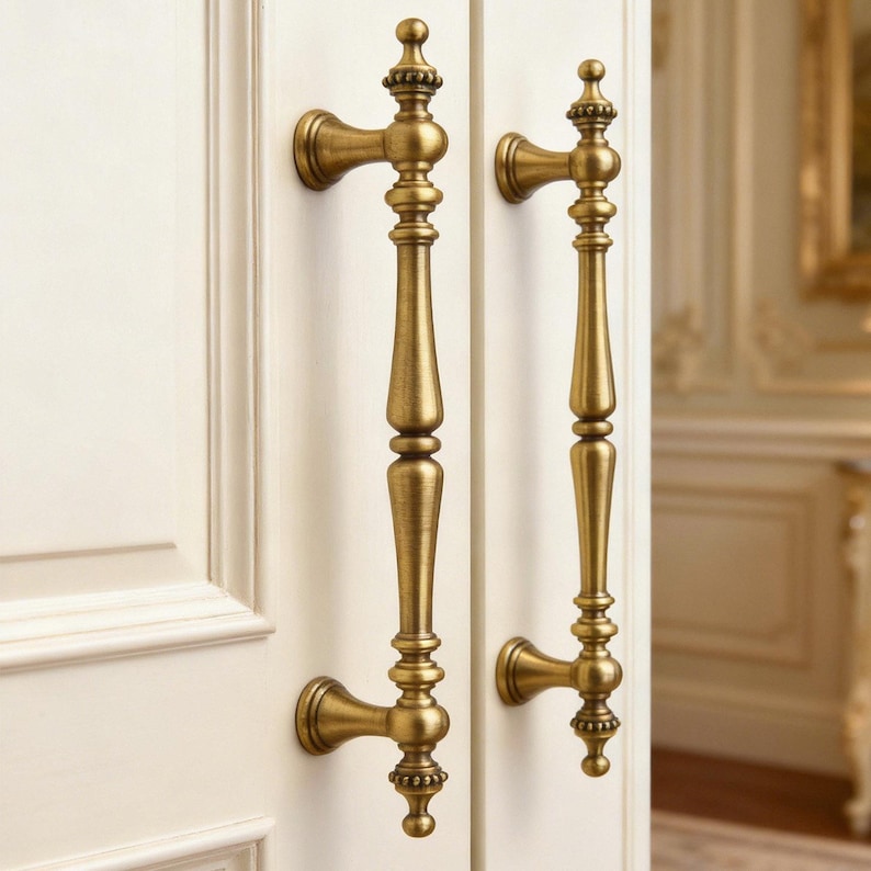 May include: Two antique brass door handles with ornate detailing. The handles are mounted on a white door, showcasing a classic design. The handles have a vertical bar with decorative elements and a curved base.