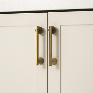 May include: Close-up of two cream-colored cabinet doors with vertical brass-colored handles. The handles are cylindrical with rounded ends. The text "ALL FOR YOUR SIMPLE LIFE" is at the bottom of the image.