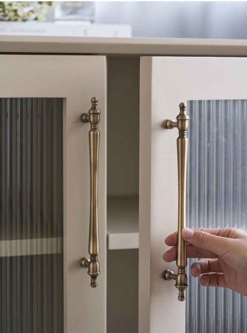 以下が含まれることがあります： 2つのドアが付いたキャビネットのクローズアップ。各ドアには、長いアンティーク真鍮のハンドルが付いています。キャビネットのドアは明るいベージュ色で、ガラスパネルはリブ加工が施されています。キャビネットは開いており、白い内部が見えます。