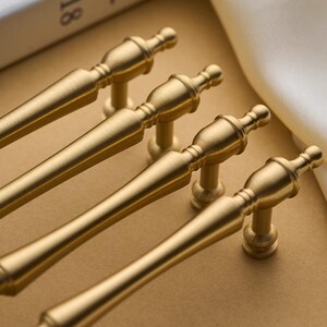 以下が含まれることがあります： クラシックなデザインの、金色のキャビネットプルが4つ。各プルは、装飾的なディテールと小さな丸いベースを備えた円筒形のハンドルを備えています。プルはニュートラルなトーンの表面に配置されており、ハードウェアまたは家の装飾のテーマを示唆しています。