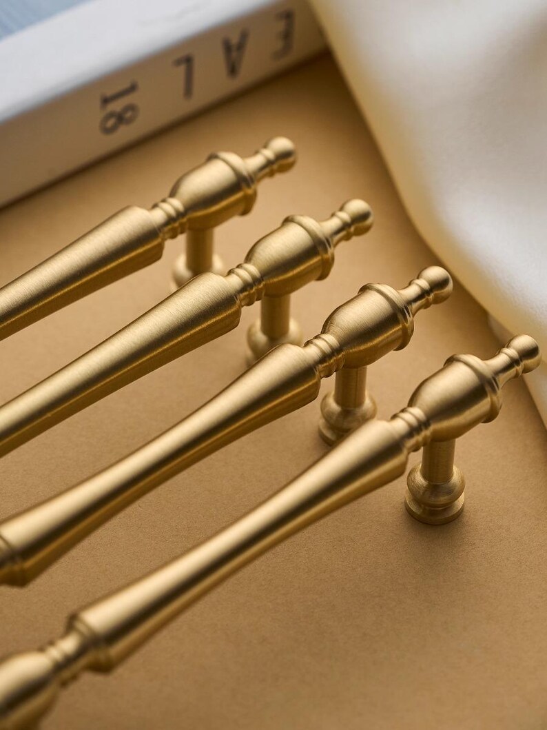 以下が含まれることがあります： クラシックなデザインの、金色のキャビネットプルが4つ。各プルは、装飾的なディテールと小さな丸いベースを備えた円筒形のハンドルを備えています。プルはニュートラルなトーンの表面に配置されており、ハードウェアまたは家の装飾のテーマを示唆しています。