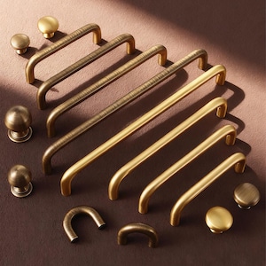 Puede incluir: Una variedad de herrajes de latón para gabinetes, que incluyen manijas y perillas, en varios tamaños y acabados. El hardware está dispuesto sobre una superficie marrón, mostrando diferentes estilos y texturas. Las manijas son curvas y las perillas son redondas.