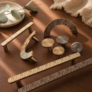 Puede incluir: Una colección de tiradores y pomos de latón con superficies texturizadas. El hardware, en varias formas y tamaños, tiene un tono dorado cálido. La disposición se encuentra sobre una superficie de madera.