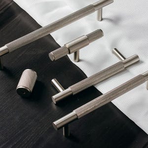 Op de afbeelding: Een verzameling zilverkleurige kastbeslagen, waaronder handgrepen en een cilindrische knop. De hardware heeft een getextureerde, diamantvormige grip. De items zijn gerangschikt op een zwart-witte ondergrond, wat wijst op een moderne designesthetiek.