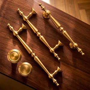 Op de afbeelding: Vier goudkleurige kastgrepen en twee bijpassende knoppen zijn gerangschikt op een donker houten oppervlak. De handgrepen hebben een sierlijk, vintage ontwerp met decoratieve details. De knoppen zijn rond en hebben een geborstelde afwerking.