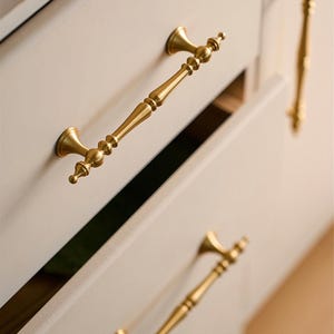 May include: Close-up of gold-colored cabinet hardware. The image shows several ornate drawer pulls and a cabinet knob. The pulls have a detailed, vintage design, with a central handle and decorative end pieces. The cabinet is painted a light neutral color.