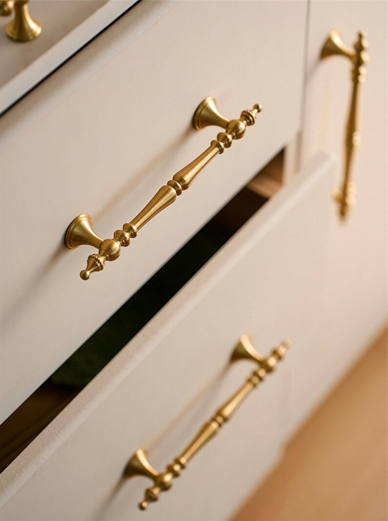 May include: Close-up of gold-colored cabinet hardware. The image shows several ornate drawer pulls and a cabinet knob. The pulls have a detailed, vintage design, with a central handle and decorative end pieces. The cabinet is painted a light neutral color.