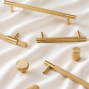 May include: A collection of gold-toned cabinet hardware, including handles and knobs. The hardware features a textured, diamond-patterned grip and a smooth, solid base. The pieces are arranged on a white, textured surface.