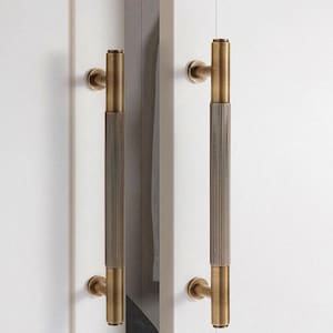 Op de afbeelding: Twee bronzen deurklinken met een verticaal, geribbeld ontwerp. De handgrepen zijn gemonteerd op een witte deur. De handgrepen hebben een cilindrische vorm met een decoratief, getextureerd patroon.