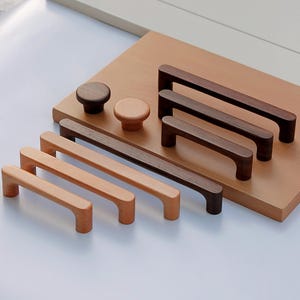 May include: A collection of wooden cabinet hardware, including knobs and handles, in various sizes and shades of brown. The hardware is displayed on a light brown wooden board, showcasing the different styles and finishes available for home decor projects.