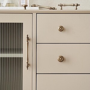 以下が含まれることがあります： アンティーク真鍮の金具が付いた、クリーム色のキャビネットのクローズアップ。キャビネットは、縦のラインが入ったガラスドア、引き出しの取っ手、丸いノブが特徴です。「Design copyright servant brand」のテキストが表示されています。