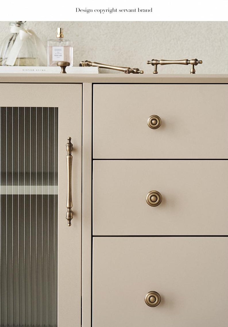 以下が含まれることがあります： アンティーク真鍮の金具が付いた、クリーム色のキャビネットのクローズアップ。キャビネットは、縦のラインが入ったガラスドア、引き出しの取っ手、丸いノブが特徴です。「Design copyright servant brand」のテキストが表示されています。
