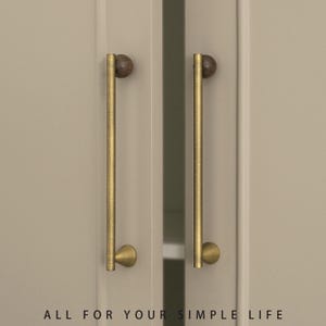 May include: Two cabinet door pulls with a brass finish and a dark wood ball at the top. The pulls are mounted on a light beige cabinet door. The text "ALL FOR YOUR SIMPLE LIFE" is at the bottom.