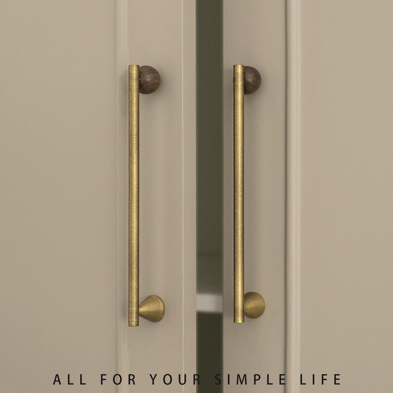 May include: Two cabinet door pulls with a brass finish and a dark wood ball at the top. The pulls are mounted on a light beige cabinet door. The text "ALL FOR YOUR SIMPLE LIFE" is at the bottom.