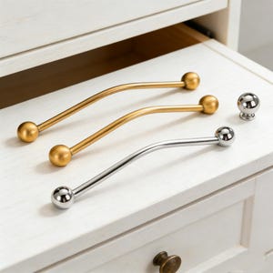 以下が含まれることがあります： 金色の曲線的なハンドル2つと銀色のハンドル1つを含む、キャビネットハードウェアのコレクション。それぞれ球状の端が付いています。お揃いのシルバーノブも付いています。ハードウェアは白い塗装面に表示されています。