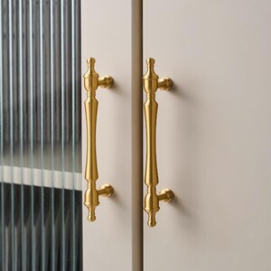 以下が含まれることがあります： 2つの金色のキャビネットドアハンドルをクローズアップ。ハンドルは垂直方向に配置され、長くスリムなデザインと装飾的なディテールが施されています。ハンドルは明るい色のキャビネットドアに取り付けられています。