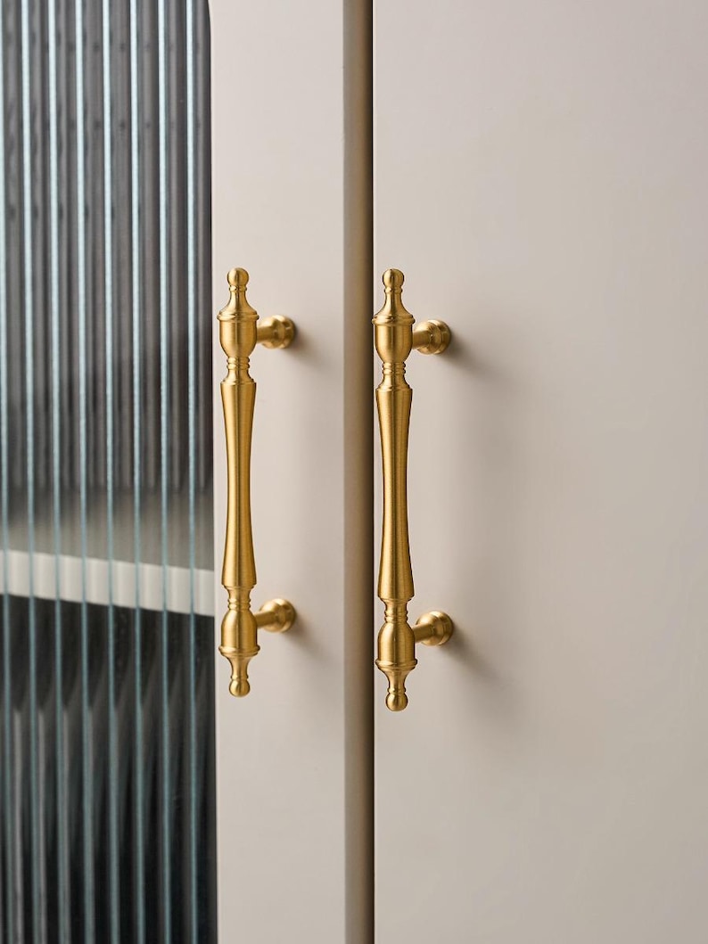 以下が含まれることがあります： 2つの金色のキャビネットドアハンドルをクローズアップ。ハンドルは垂直方向に配置され、長くスリムなデザインと装飾的なディテールが施されています。ハンドルは明るい色のキャビネットドアに取り付けられています。