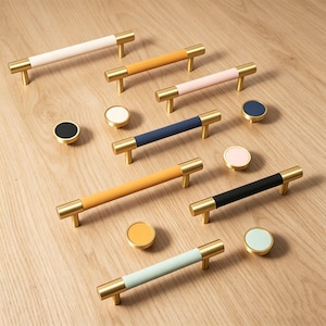 Può includere: Una collezione di ferramenta per mobili, tra cui maniglie dorate con impugnature in pelle colorata bianca, arancione, rosa, blu navy, gialla e nera. Sono presenti anche pomelli rotondi coordinati nei colori.
