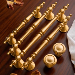 Könnte beinhalten: Eine Sammlung goldfarbener Schrankbeschläge, darunter vier lange Griffe und zwei runde Knöpfe. Die Griffe haben ein detailliertes, verziertes Design, und die Knöpfe weisen einen perlenbesetzten Rand auf. Die Hardware wird auf einer dunklen Holzoberfläche präsentiert.