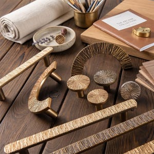 Può includere: Una collezione di ferramenta in ottone, tra cui maniglie e pomelli, con finitura strutturata. L'hardware ha una calda tonalità dorata. Altri oggetti includono un taccuino, pennelli e un piattino. Misure in cm.