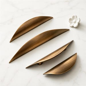 Puede incluir: Cuatro tiradores de gabinete de color bronce y un pomo blanco de cerámica en forma de flor están dispuestos sobre una superficie de mármol blanco. Los tiradores tienen un diseño curvo en forma de hoja. La ferretería es probablemente para muebles o gabinetes.