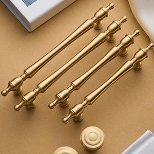 May include: A collection of gold-colored cabinet hardware, including four different-sized pulls and two round knobs. The pulls have a classic, ornate design with decorative ends. The hardware is arranged on a neutral-toned surface.