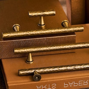 May include: Close-up of several antique brass cabinet pulls and knobs with a hammered texture. The hardware is displayed on a brown surface, showcasing the warm, metallic finish and decorative design. The pulls are in various shapes.