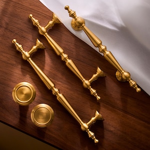 Op de afbeelding: Een verzameling goudkleurige kastbeslagen, waaronder twee knoppen en drie handgrepen. De hardware is gerangschikt op een donker houten oppervlak met witte stof op de achtergrond. De handgrepen hebben een sierlijk, vintage ontwerp.