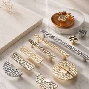 May include: A selection of decorative cabinet hardware in gold, silver, and bronze hues. The collection features various handle styles, knobs, and a decorative tray, all with a textured, hammered metal finish. The hardware is displayed on a marble surface.