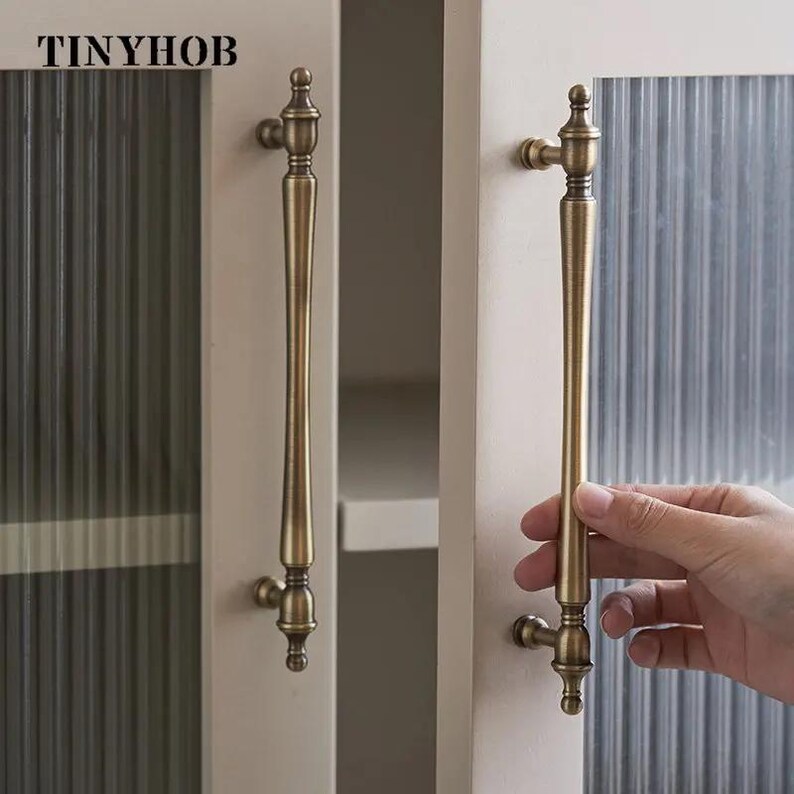 以下が含まれることがあります： 縦長のアンティーク真鍮製キャビネットハンドルが2つ。ハンドルは、テクスチャードガラスパネルが付いた白いキャビネットドアに取り付けられています。「TINYHOB」の文字が上部に印刷されています。