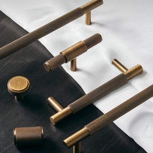 May include: A collection of bronze-colored cabinet hardware, including knobs and bar pulls. The hardware features a textured, diamond-patterned grip and a smooth, cylindrical base. The items are arranged on a black and white surface, showcasing their design and finish.
