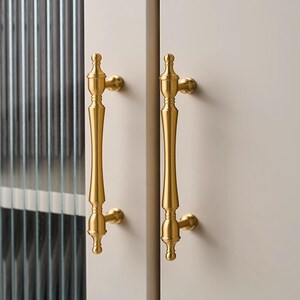 以下が含まれることがあります： 2つの金色のキャビネットドアハンドル。各ハンドルは、上部と下部に装飾的なフィニアルが付いた、長く細いボディを備えています。ハンドルは明るい色のキャビネットドアに取り付けられており、左側にはテクスチャードガラスパネルが見えます。