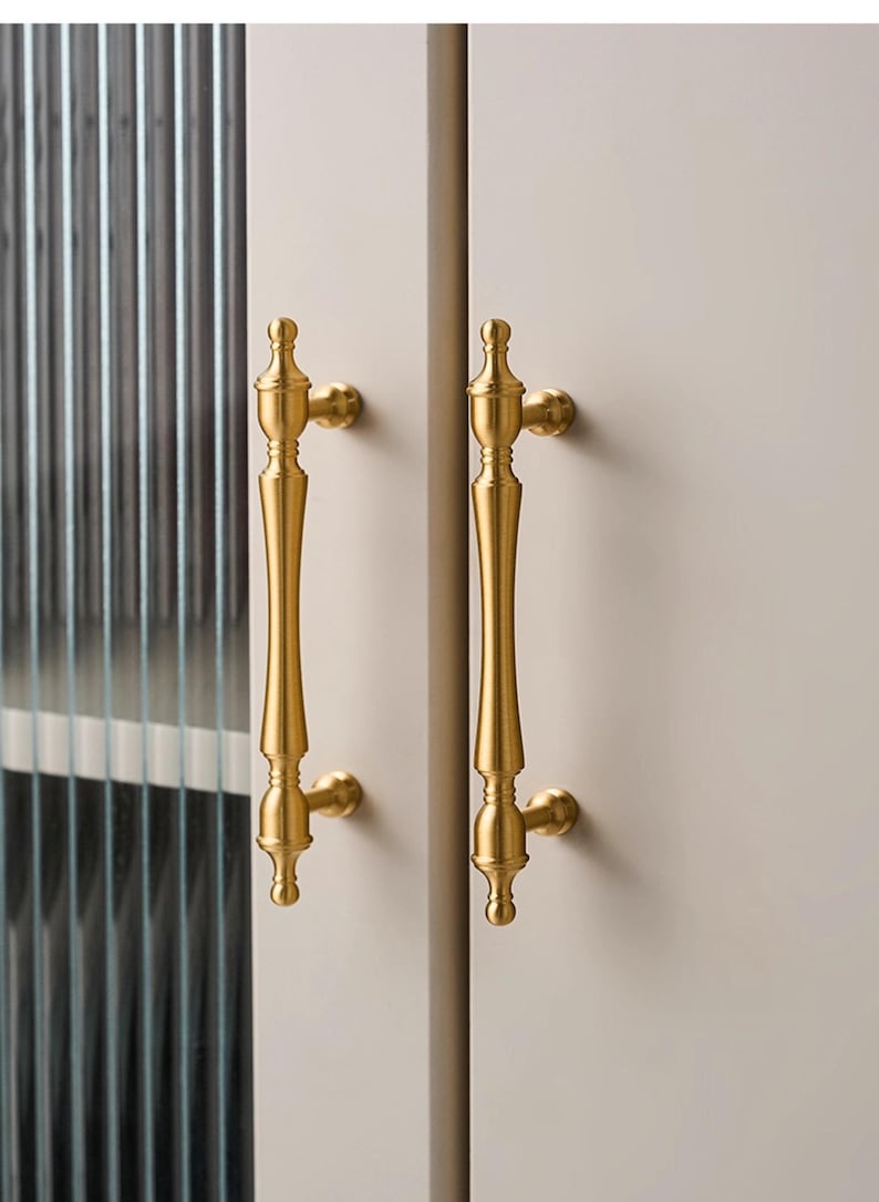 以下が含まれることがあります： 2つの金色のキャビネットドアハンドル。各ハンドルは、上部と下部に装飾的なフィニアルが付いた、長く細いボディを備えています。ハンドルは明るい色のキャビネットドアに取り付けられており、左側にはテクスチャードガラスパネルが見えます。