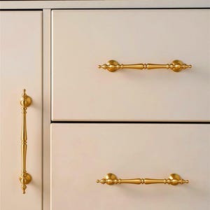 May include: Close-up of a cream-colored cabinet with gold-colored hardware. The cabinet has three drawers and a door, each with a different style of gold handle. The top of the cabinet has a small gold handle and a book with the text "Collection 2023".