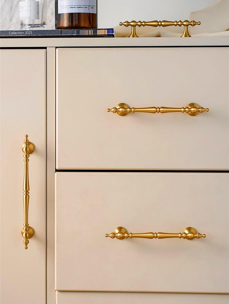 May include: Close-up of a cream-colored cabinet with gold-colored hardware. The cabinet has three drawers and a door, each with a different style of gold handle. The top of the cabinet has a small gold handle and a book with the text "Collection 2023".
