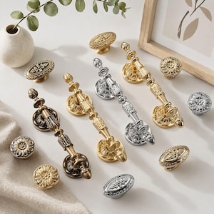 May include: A selection of decorative cabinet hardware, including knobs and pulls, in gold and silver finishes. The hardware features ornate, vintage-inspired designs with floral and scrollwork details. The knobs and pulls are arranged on a white surface.