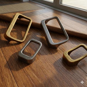 May include: Four metal cabinet pulls in varying shades of bronze and silver are arranged on a wooden surface. The pulls have a rectangular design with a curved base, suggesting a modern or industrial aesthetic. The lighting highlights the metallic textures.